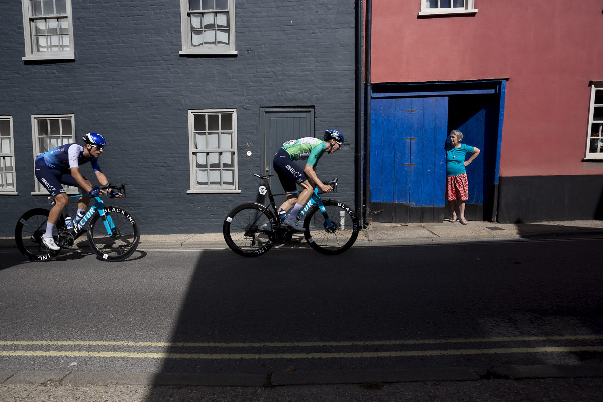 Tour of Britain 2024 - Stevie Williams and Ethan Vernon of Israel - Premier Tech pass by cottages while a women views them from her doorway