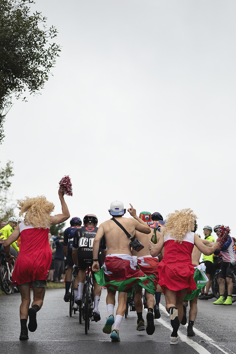 Tour of Britain 2025 - A group of stragglers are chased up the hill by men dressed as cheerleaders and topless men wearing welsh flags round their waists