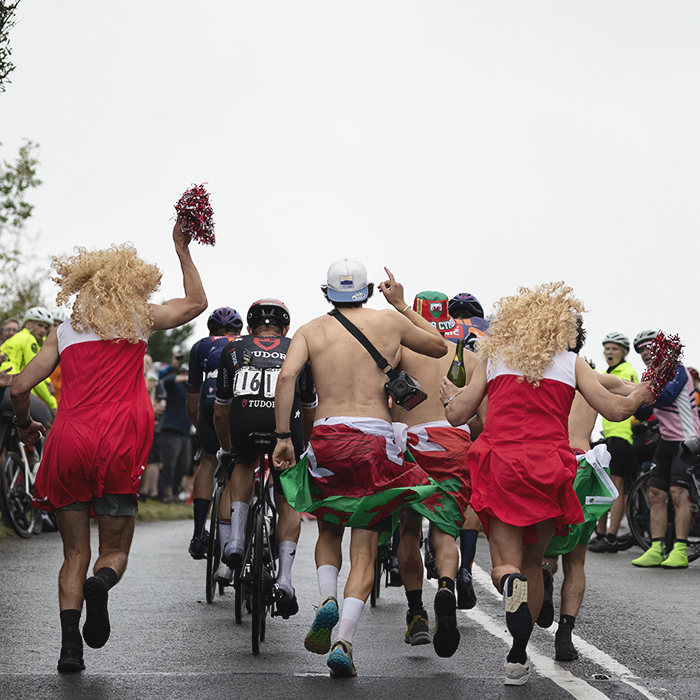 Tour of Britain 2025 - A group of stragglers are chased up the hill by men dressed as cheerleaders and topless men wearing welsh flags round their waists
