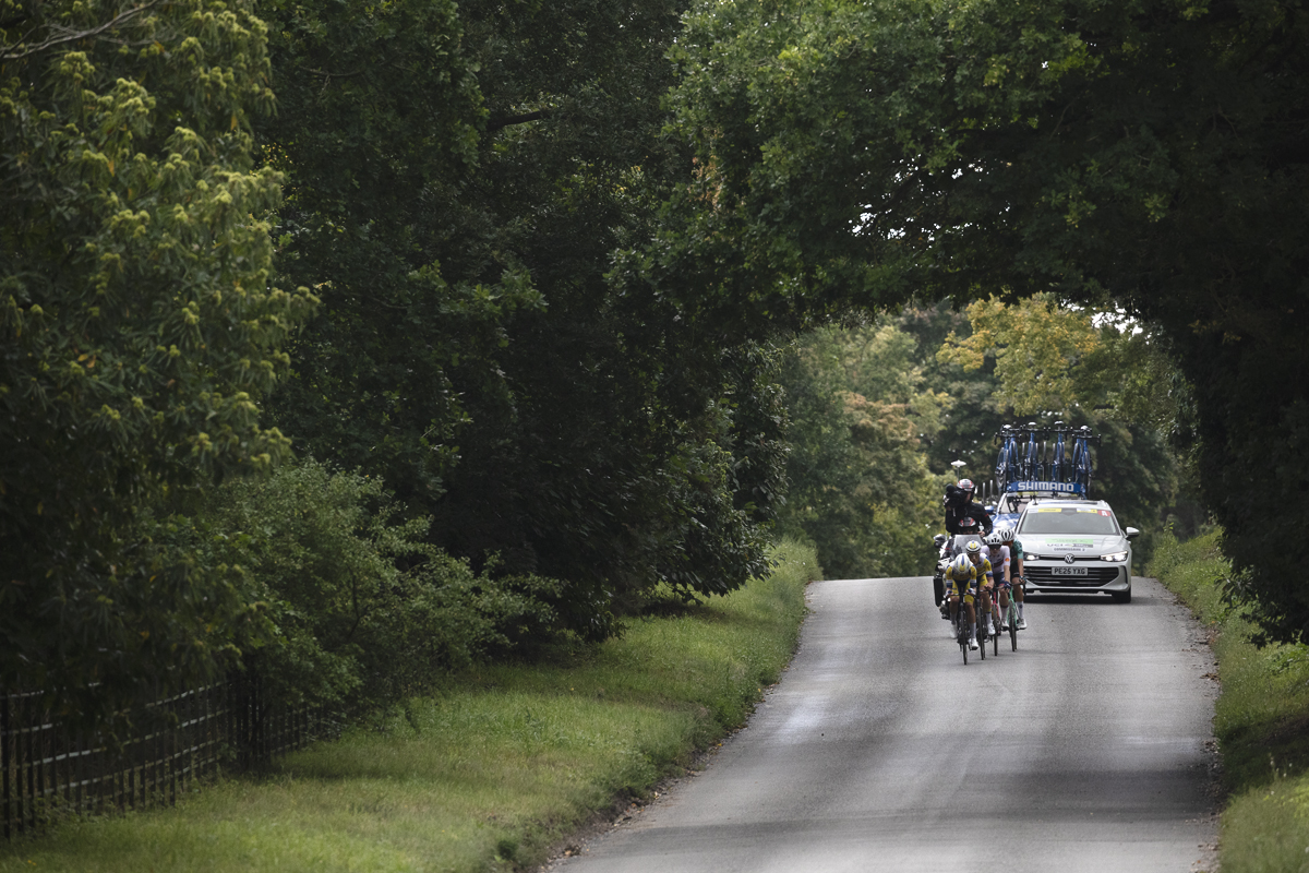Tour of Britain 2025 - Trees create an archway over the road framing the riders as they pass underneath