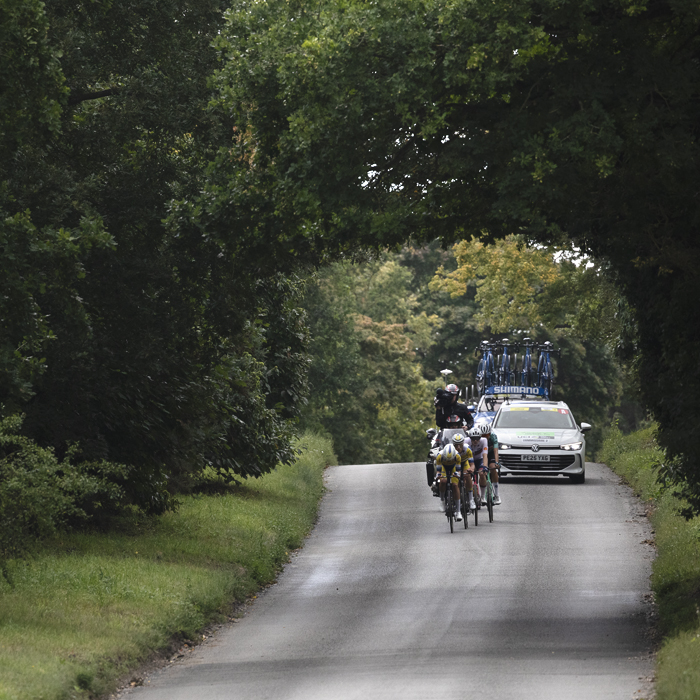 Tour of Britain 2025 - Trees create an archway over the road framing the riders as they pass underneath