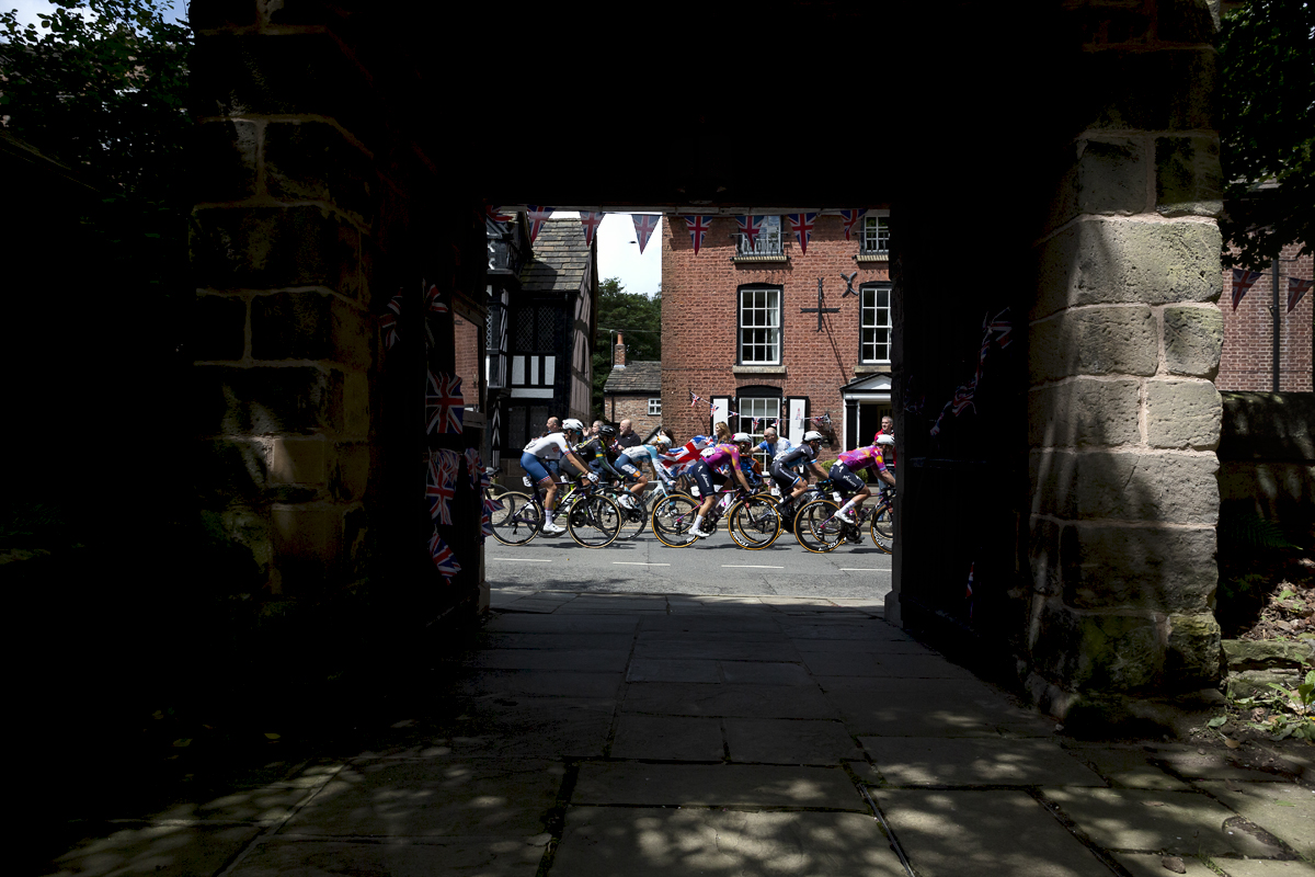 Tour of Britain Women’s 2024 - The riders race by flag waving fans framed by the church gate in The Village