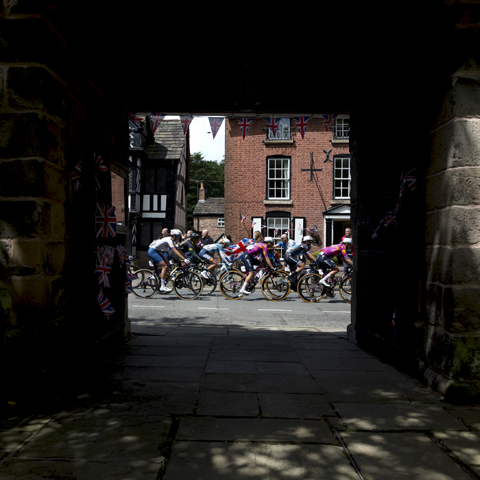 Tour of Britain Women’s 2024 - The riders race by flag waving fans framed by the church gate in The Village