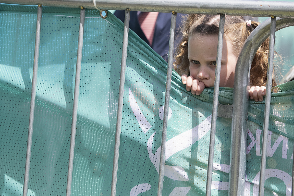 Tour of Britain Women’s 2024 - A young girl pulls down the edge of a banner so that she can see through the railings to the riders