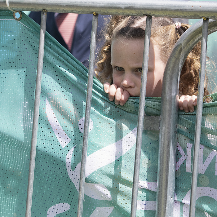 Tour of Britain Women’s 2024 - A young girl pulls down the edge of a banner so that she can see through the railings to the riders