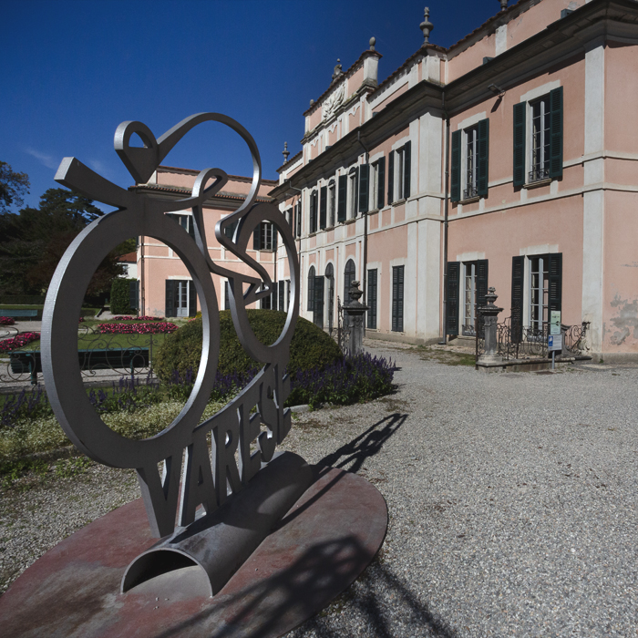 Tre Valli Varesine 2025 - A statute of a cyclist in a tuck position crafted from metal stands in the courtyard of a grand pink coloured building in Varese