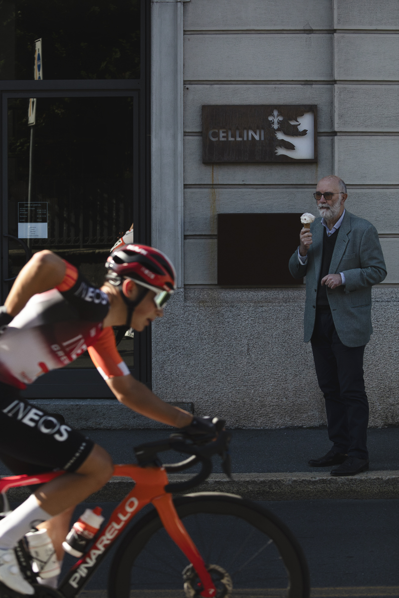 Tre Valli Varesine 2025 - A man with a beard eats a gelato while watching a rider pass by who is reaching for a gel