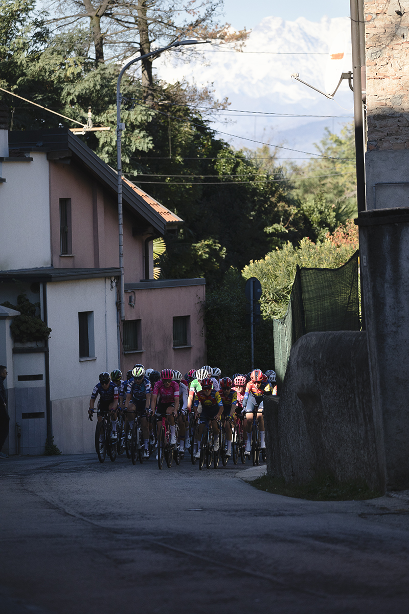 Tre Valli Varesine Women’s Race 2025 - The peloton passes through a narrow street with the snow capped Monte Rossa in the background