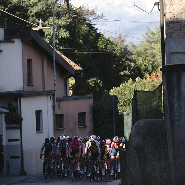 Tre Valli Varesine Women’s Race 2025 - The peloton passes through a narrow street with the snow capped Monte Rossa in the background
