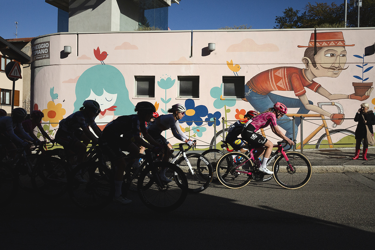 Tre Valli Varesine Women’s Race 2025 - Mirre Knaven of EF Education-Oatly leads a group of riders out of the shadows past a cheerful mural of a man in a hat holding a plant whilst riding a bike