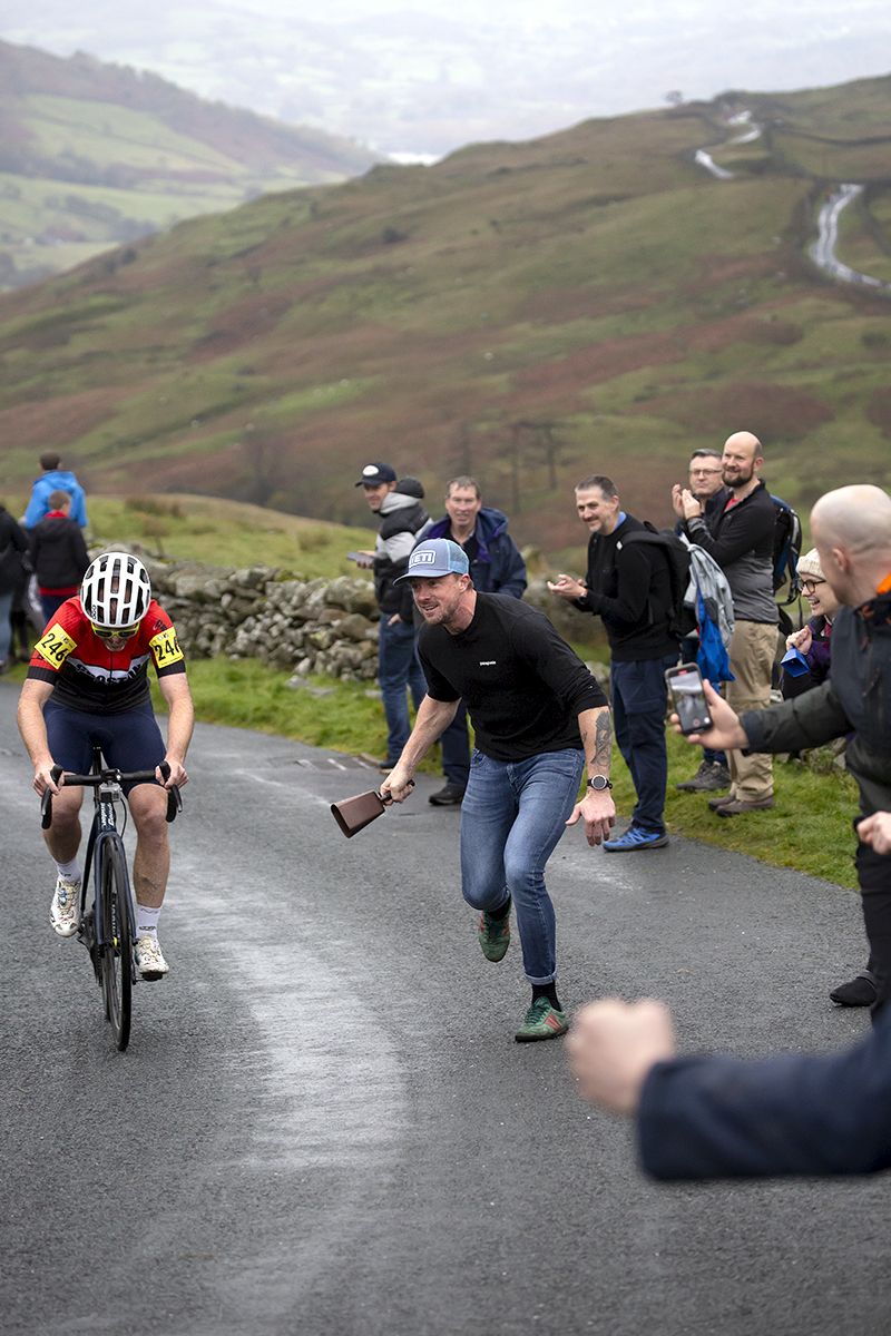 UK National Hill Climb Championships 2023 - A fan with a large cowbell runs alongside Chris Breen