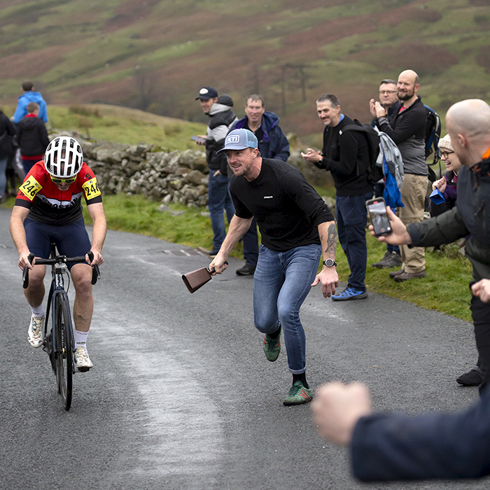 UK National Hill Climb Championships 2023 - A fan with a large cowbell runs alongside Chris Breen